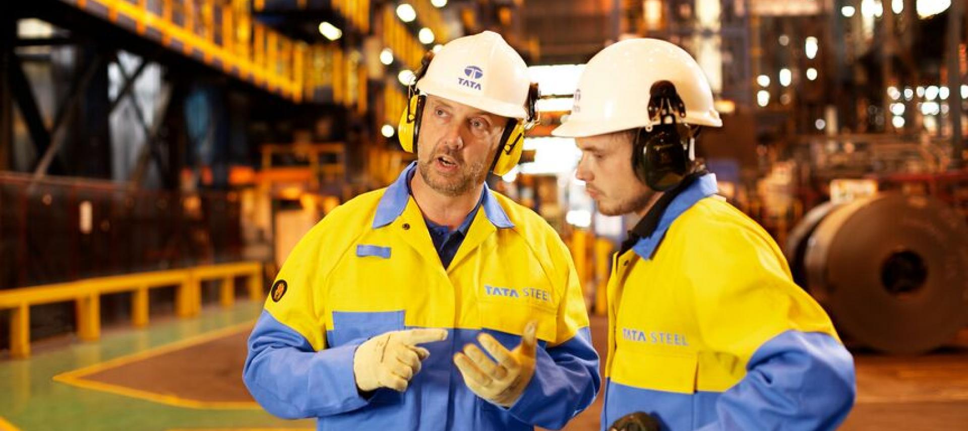 Tata Steel workers in a manufacturing environment, wearing protective equipment, having a discussion